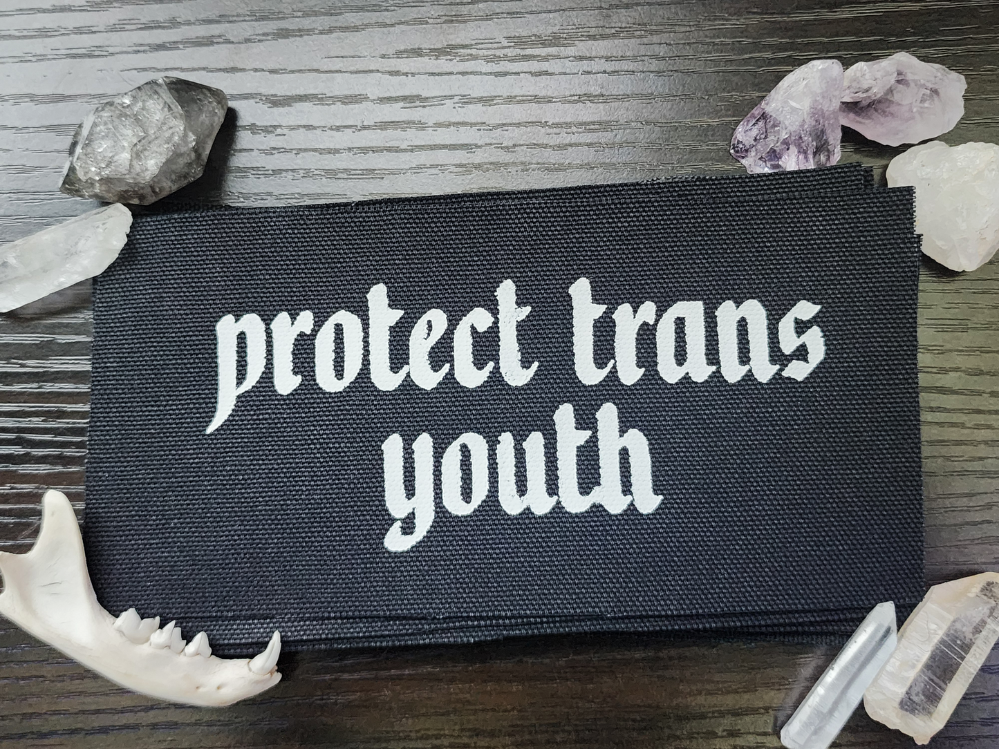 A black fabric patch with the phrase 'Protect Trans Youth' printed in bold, white Gothic-style lettering. The patch is stacked neatly with others on a dark wooden surface. Surrounding the patch are decorative elements, including clear and purple crystals, a small animal jawbone, and rough stones, adding a mystical and earthy aesthetic.