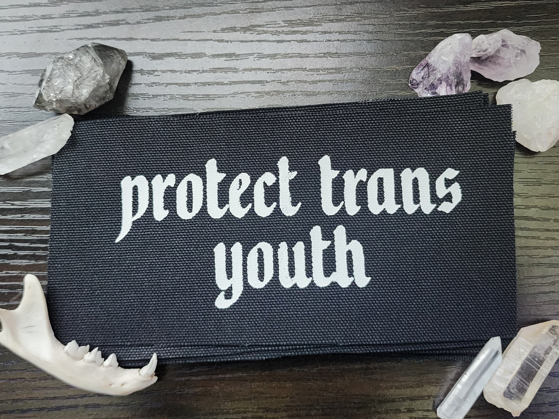A black fabric patch with the phrase 'Protect Trans Youth' printed in bold, white Gothic-style lettering. The patch is stacked neatly with others on a dark wooden surface. Surrounding the patch are decorative elements, including clear and purple crystals, a small animal jawbone, and rough stones, adding a mystical and earthy aesthetic.