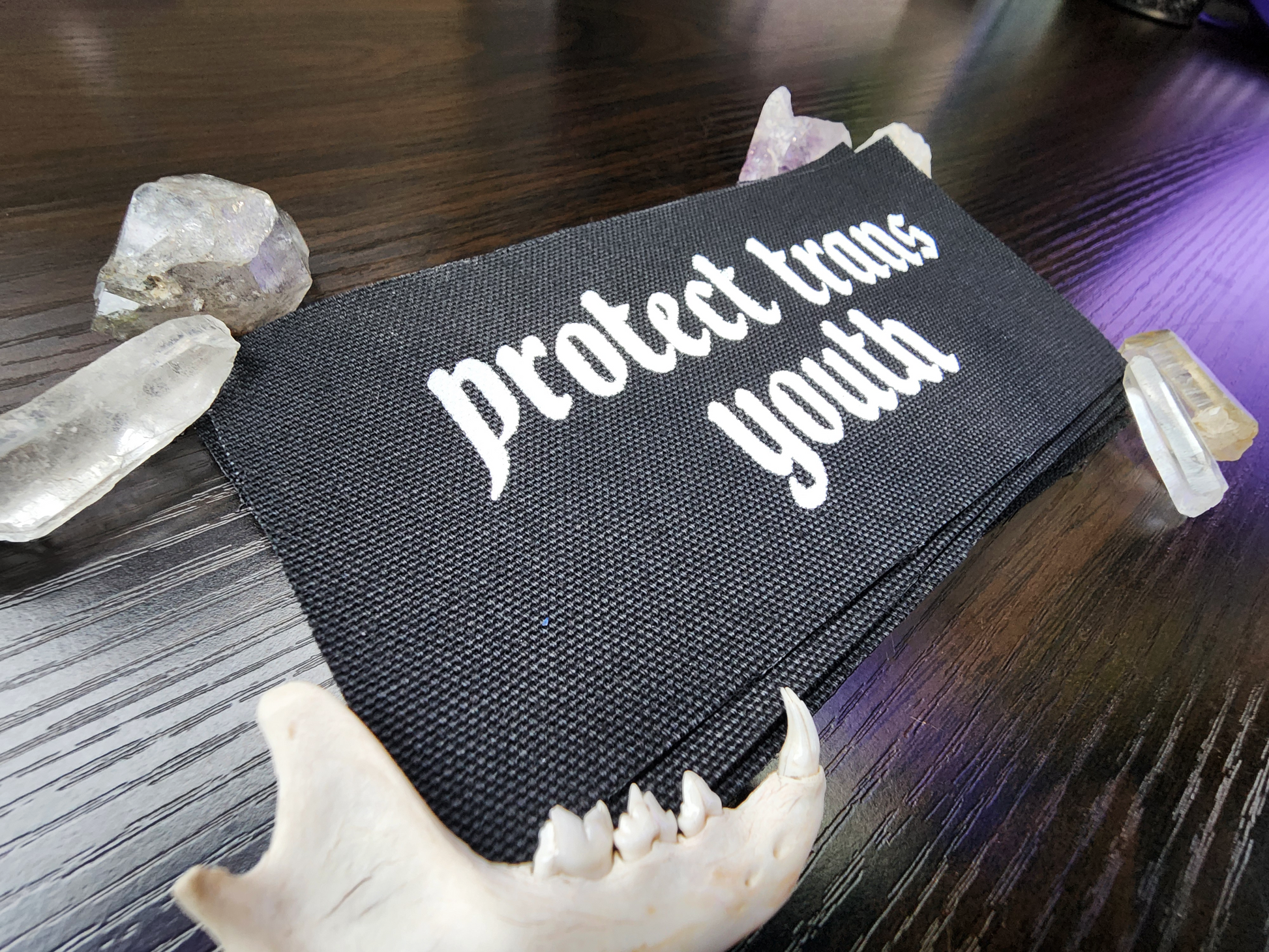 A black fabric patch with the phrase 'Protect Trans Youth' printed in bold, white Gothic-style lettering. The patch is stacked neatly with others on a dark wooden surface. Surrounding the patch are decorative elements, including clear and purple crystals, a small animal jawbone, and rough stones, adding a mystical and earthy aesthetic.